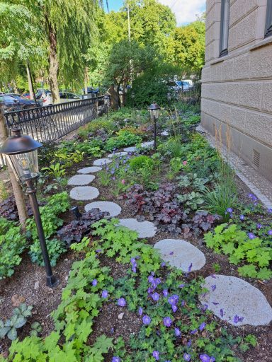 Nyplantering En grönskande trädgård med stigar av stenplattor och blommor runt en byggnad.